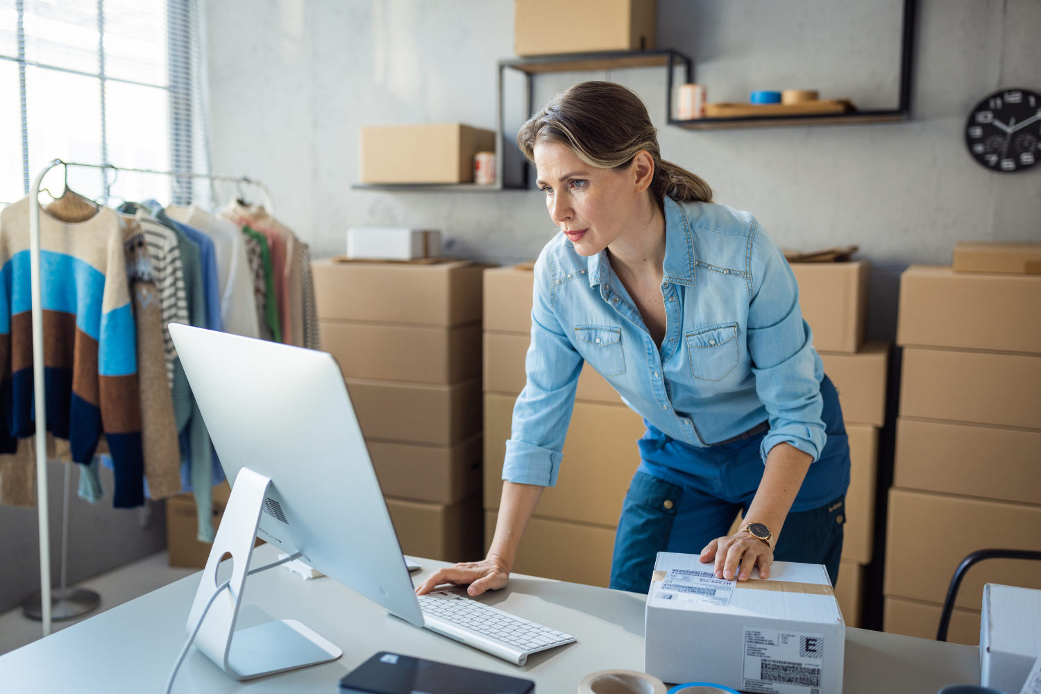 Business owner looking at laptop with boxes of orders behind her.