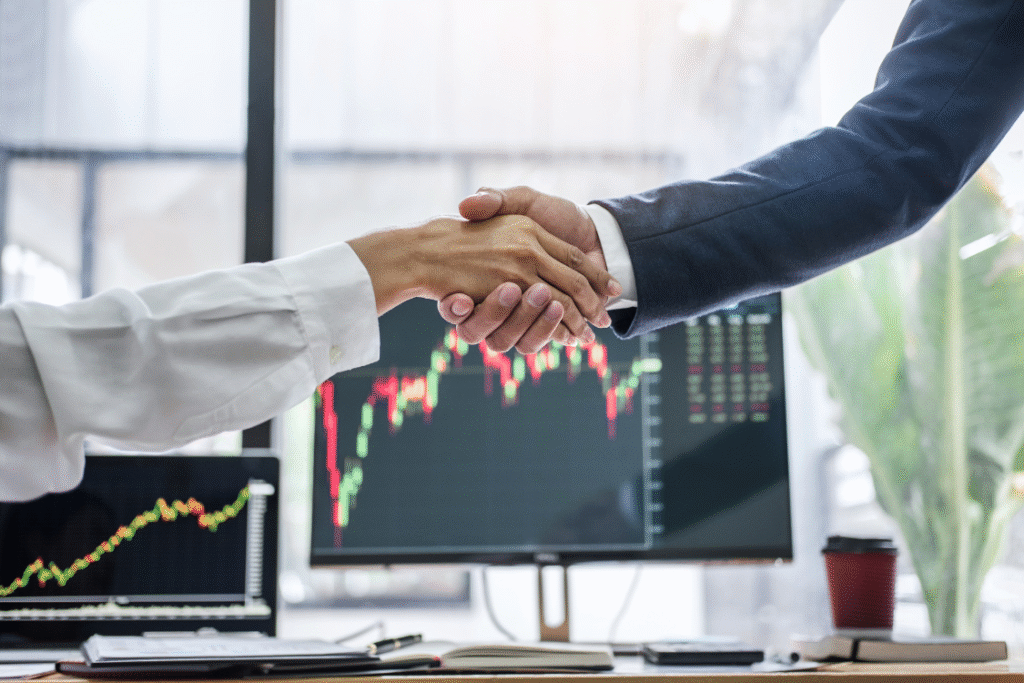 Business broker and client shaking hands in front of stock charts after closing a successful business sale