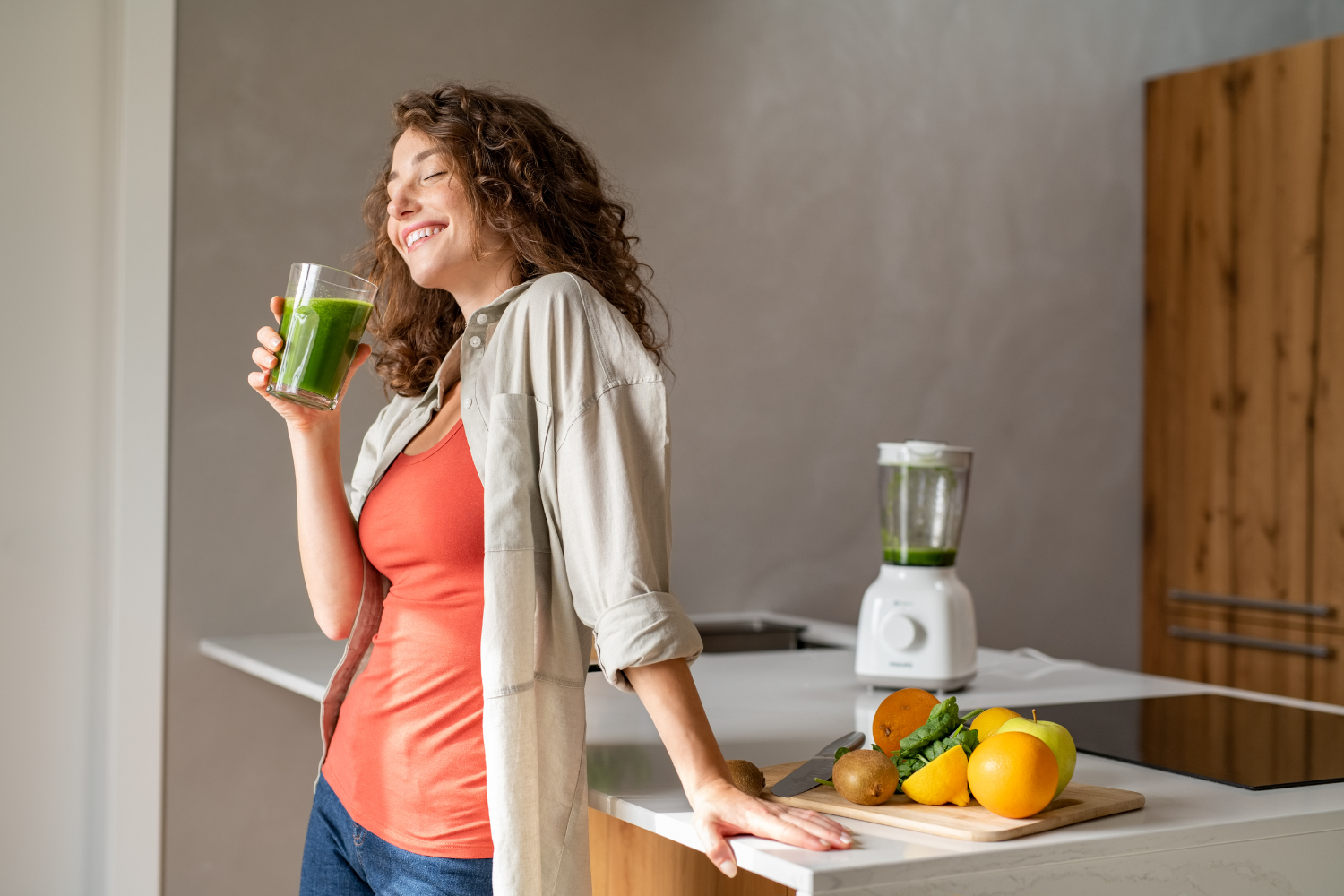 A woman drinking green detox juice at a sunny kitchen table.