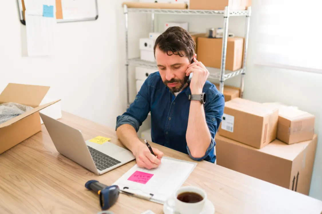 Small business owner on phone discussing supplier evaluation with notes and laptop