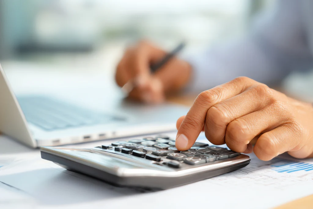 Close-up of a hand pressing keys on a calculator next to a laptop, symbolizing a business owner calculating supplement demand forecasting and financial plans.