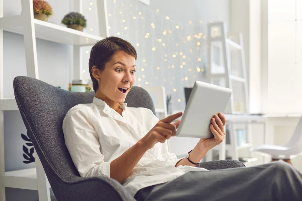 A surprised woman sitting in a chair and looking at a tablet screen, reacting positively to a major deal or a large supplement manufacturing volume discount on a bulk order.