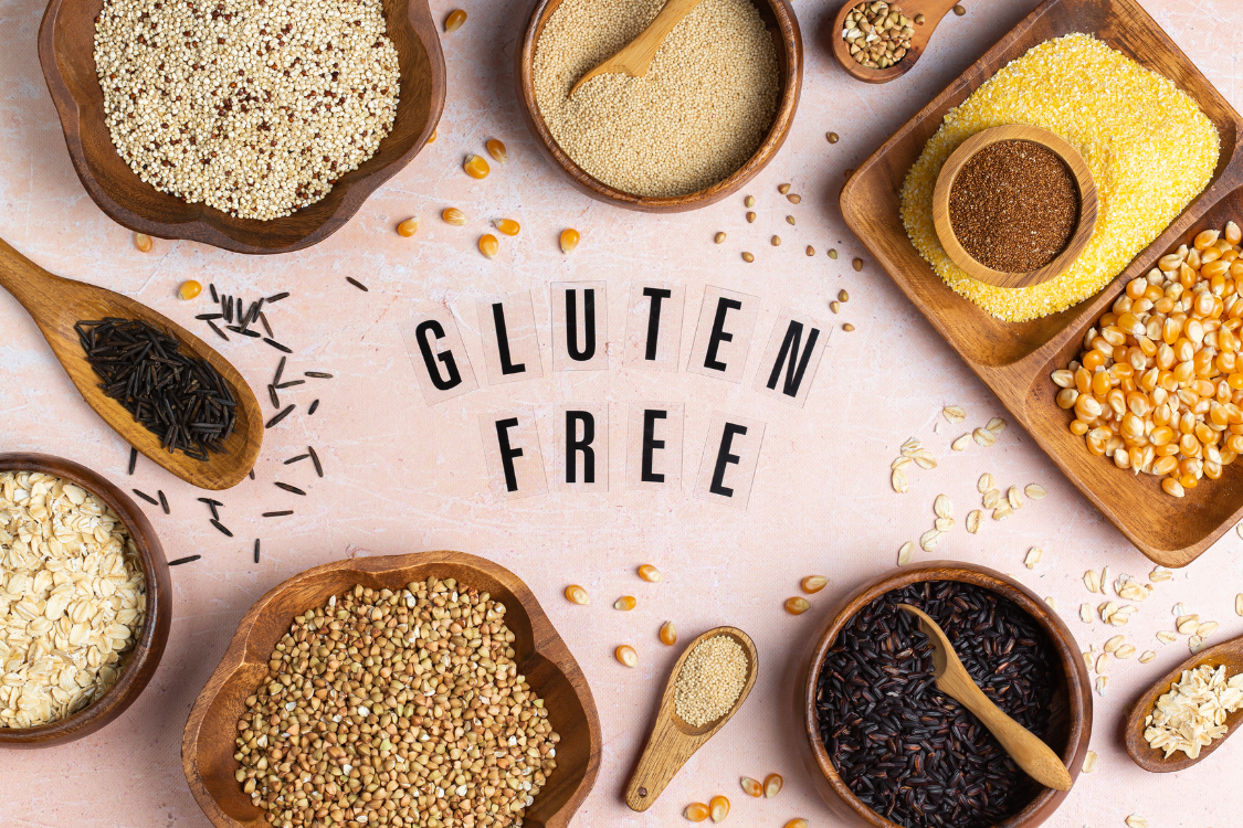 Overhead view of various gluten-free grains (quinoa, rice, corn) in wooden bowls with the words "GLUTEN FREE" spelled out in the center, illustrating ingredient control for a gluten testing supplements manufacture.