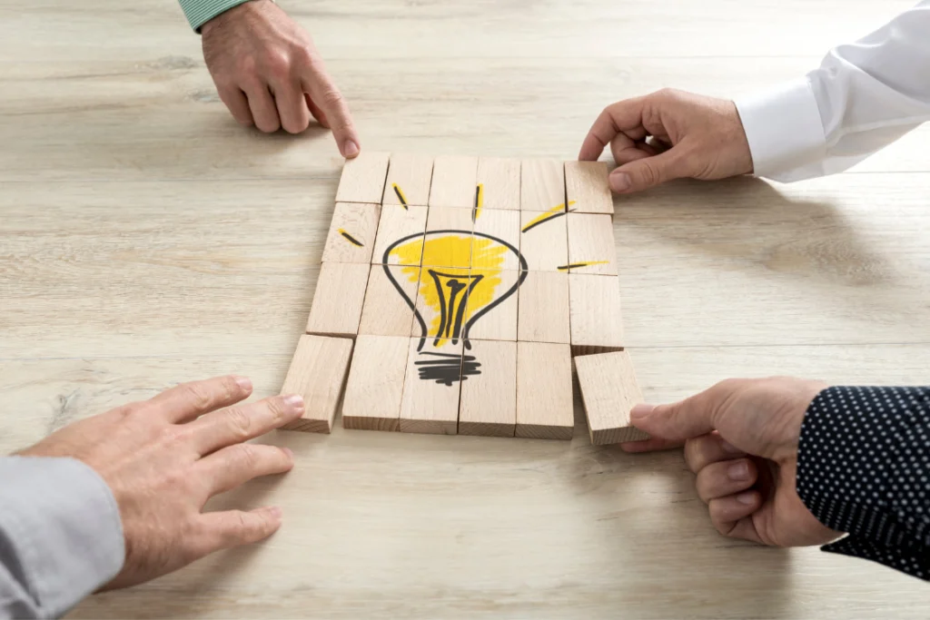 Hands assembling wooden blocks to complete a lightbulb design, symbolizing a collaborative idea or a strategy built by a brand and a low MOQ supplement manufacturer.