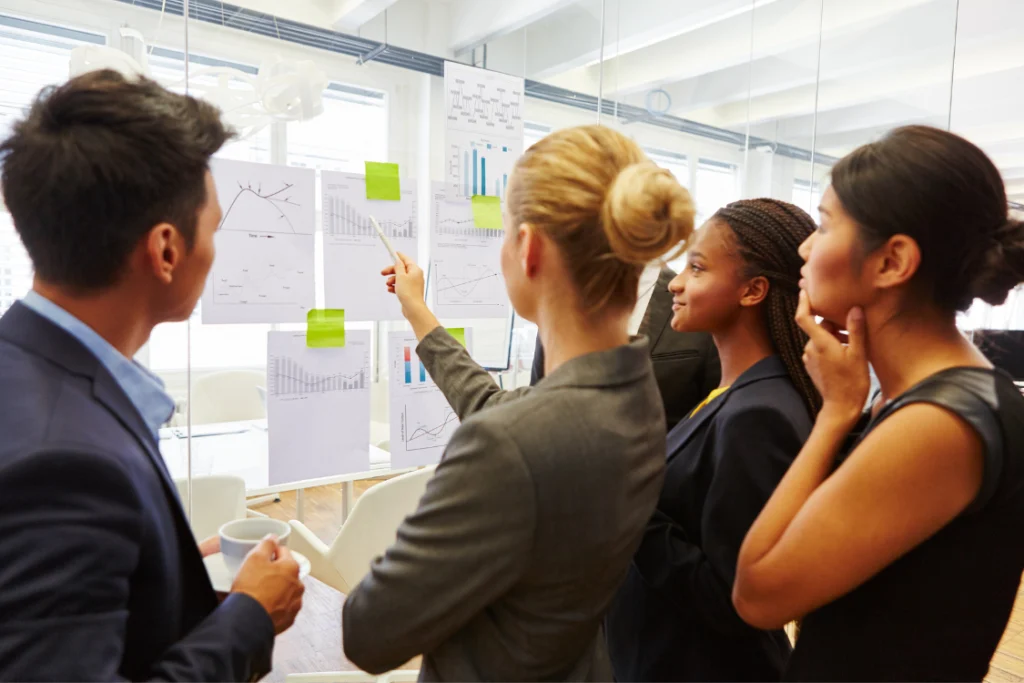 A small group of business professionals reviewing charts and graphs on a wall during a meeting, representing the review of sales data before scaling with a low MOQ supplement manufacturer.