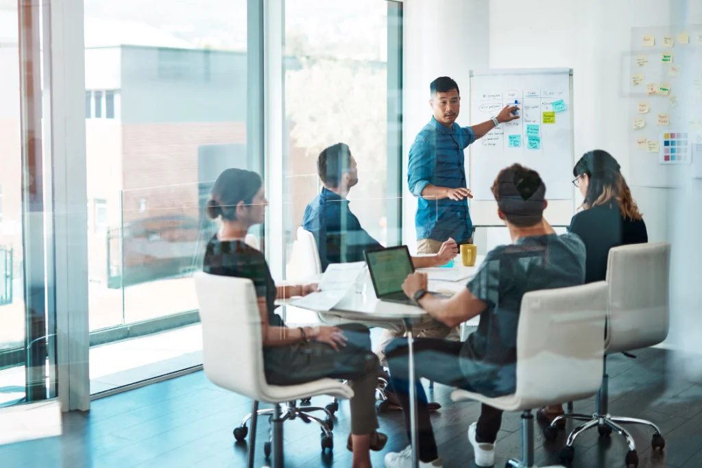 A team in a modern office, with one person presenting a plan on a whiteboard to colleagues, discussing strategy for working with a low MOQ supplement manufacturer.