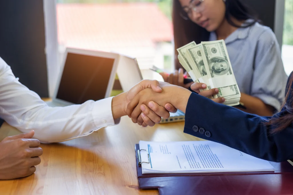 Business partners shaking hands over contract documents and money, symbolizing negotiation and cost savings in supplement manufacturer pricing comparison.