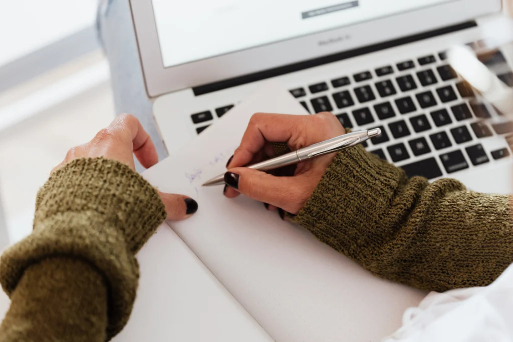 A person taking notes with a pen and notebook next to a laptop, documenting findings from supplement manufacturer public records.