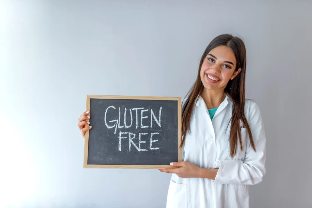 Smiling woman in a white lab coat (representing a professional in a supplements manufacture) holding a chalkboard sign that reads "GLUTEN FREE" in white chalk, emphasizing the commitment to gluten testing standards.