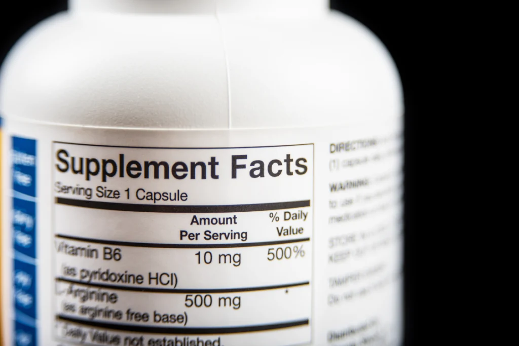 Close-up on a white supplement bottle showing the Supplement Facts label with Vitamin B6 and Arginine, emphasizing the importance of accurate labeling and FDA regulations.