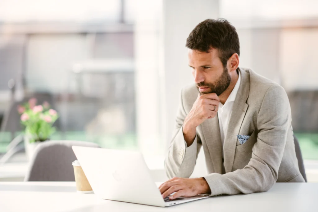 Focused businessman in a suit jacket working on a laptop in a bright modern office, performing research or online checks for a supplement manufacturing partner as part of due diligence.