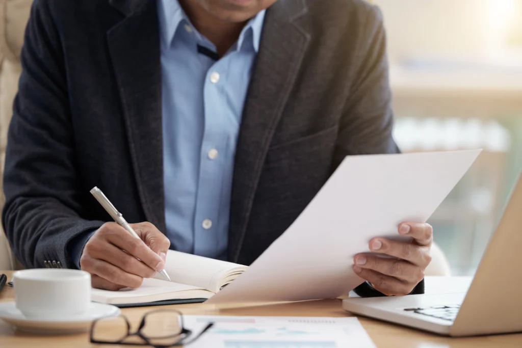 Close-up of a business executive in a suit reviewing documentation and writing notes, representing the due diligence process of checking contracts and financial stability for a supplement manufacturing partner.