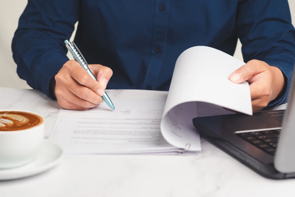 A person signing a document next to a laptop and a coffee cup, illustrating the final step of signing a supplement manufacturing agreement for cost effective and high-quality production with a proven track record.