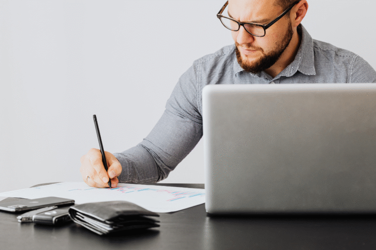 Man in business attire filling out a document with a pen next to a laptop, representing due diligence and working on a supplement manufacturer checklist.