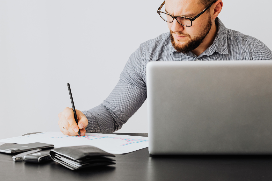 Man in business attire filling out a document with a pen next to a laptop, representing due diligence and working on a supplement manufacturer checklist.