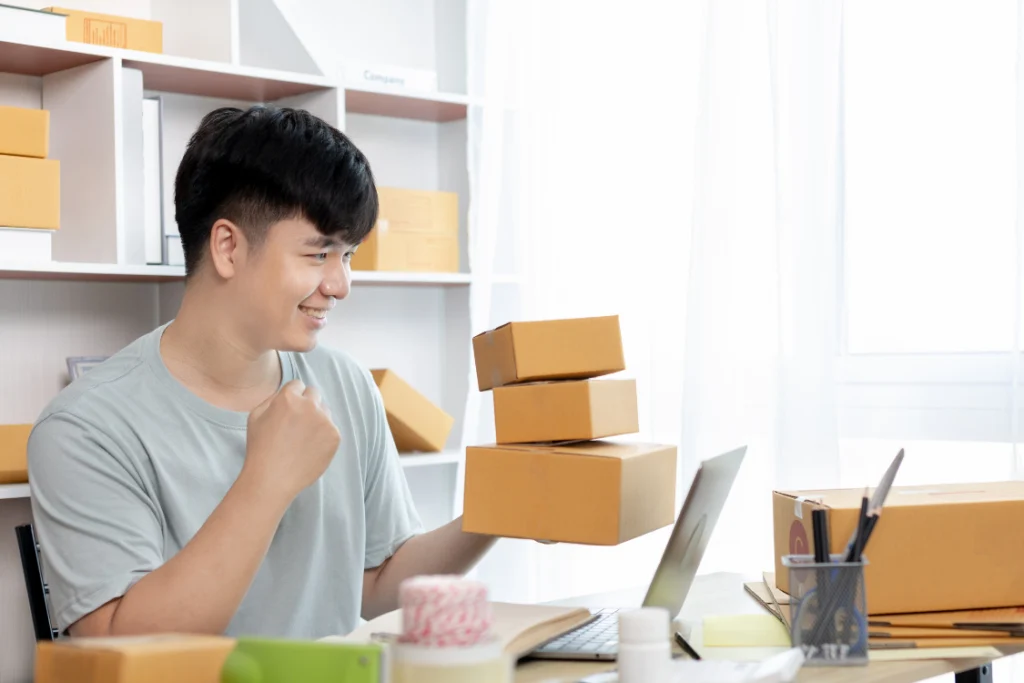 A smiling, happy dropshipper holding small, packaged boxes, celebrating a sale of private label dropshipping supplements while working on a laptop.
