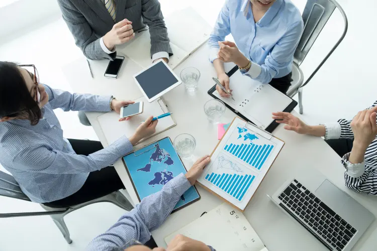 Overhead view of a business meeting with four people discussing charts and a map on a table, representing the planning stages of how to start a supplement company.