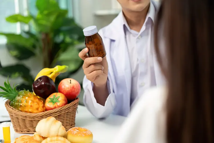 A doctor or nutritionist in a white coat holding up a bottle of supplements while sitting across from a patient, with a basket of fresh fruit in the foreground, highlighting the need for safe and informed product development.