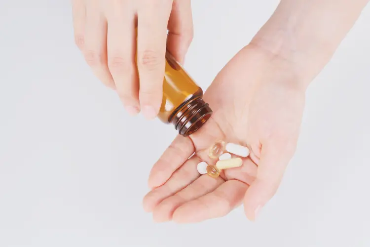 A person pouring a mixture of white, yellow, and clear softgel supplements from an amber glass bottle into the palm of their hand, representing product sampling or dosage planning.